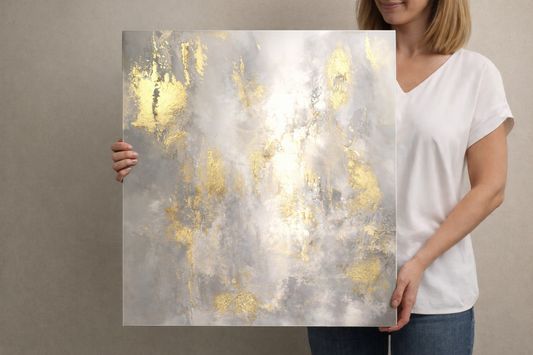 Woman holding a square abstract aluminum wall art print with soft gray and white tones and metallic gold accents, glossy non-brushed aluminum surface shown close-up against a neutral background