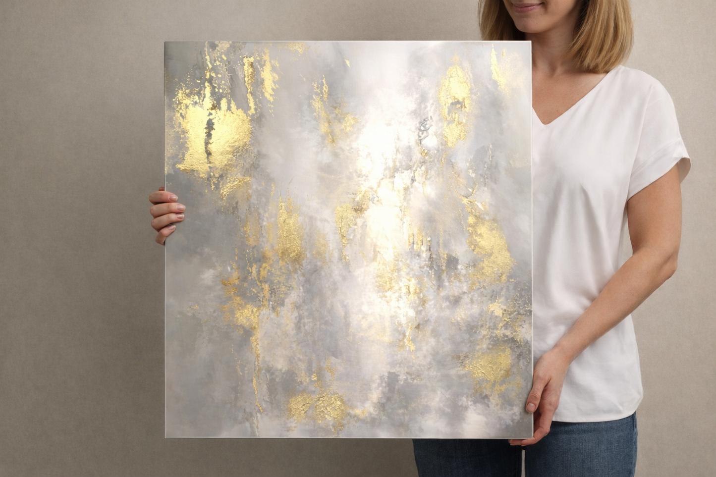 Woman holding a square abstract aluminum wall art print with soft gray and white tones and metallic gold accents, glossy non-brushed aluminum surface shown close-up against a neutral background